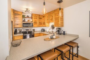 A kitchen with wooden cabinets, a granite countertop, and bar stools. - NEW! Walk to Town | Hot Tub + Game Room | Sleeps 8 (Big Sky)