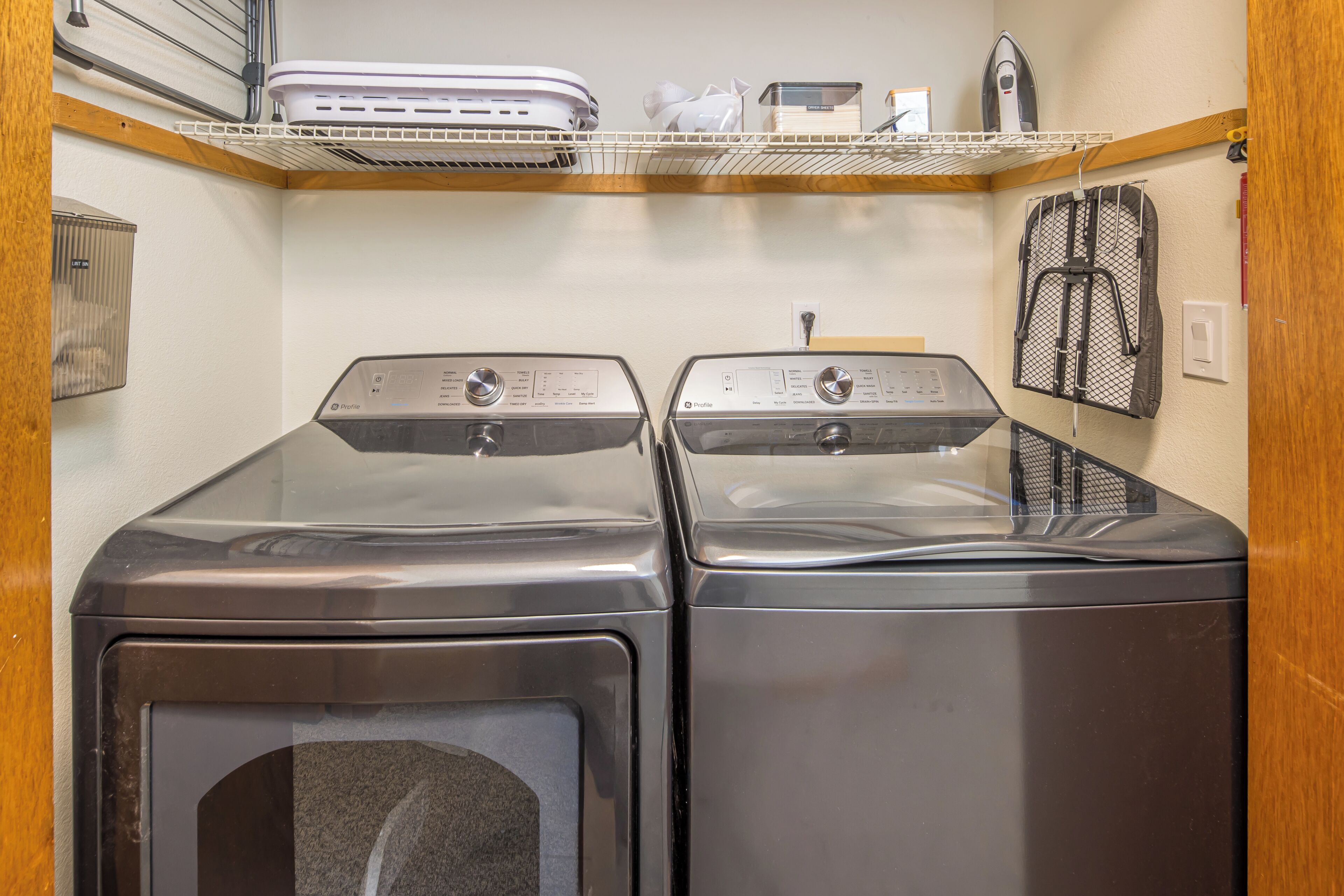 A laundry room with two front-loading washers and dryers, a shelf with a dryer, and a mesh laundry basket on the wall.