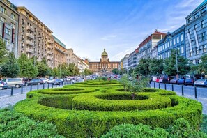 Ein Stadtplatz mit einem zentralen Garten, Gebäuden an beiden Seiten und einem markanten Gebäude mit Kuppel im Hintergrund.