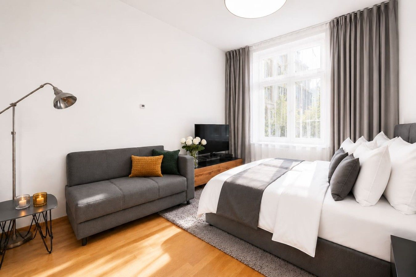 Ein modernes Schlafzimmer mit grauem Sofa, einem Bett mit weißer und grauer Bettwäsche, einem Fernseher auf einem Holzständer und einer Stehlampe.