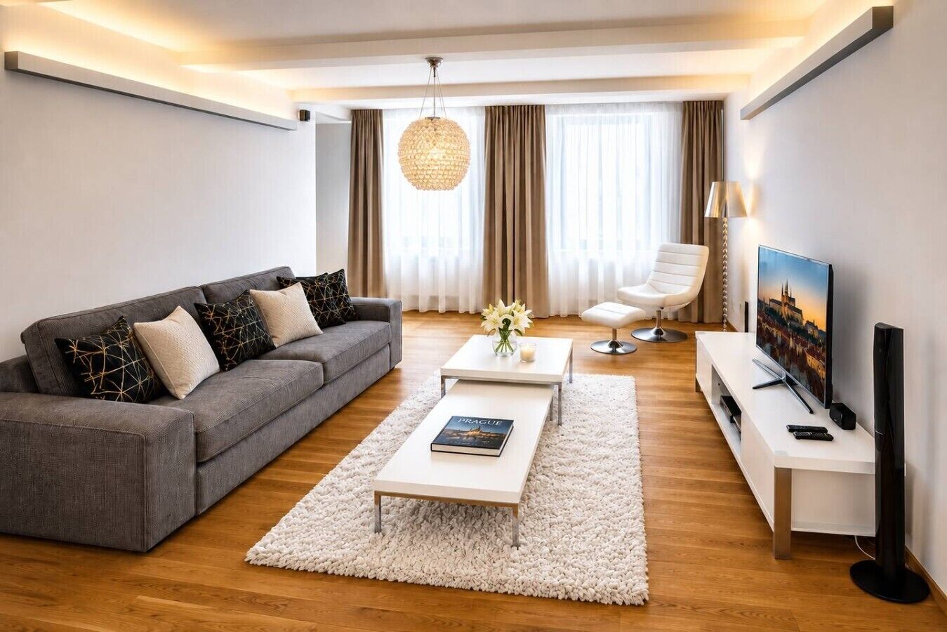 A modern living room with a grey sofa, a white coffee table, a white armchair, a TV, and a large window with curtains.