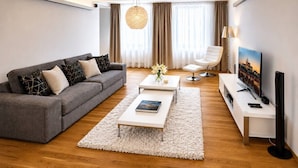 A modern living room with a grey sofa, a white coffee table, a white armchair, a TV, and a large window with curtains. - Riverside Urban Oasis in Prague’s Coolest District (Prague 8)