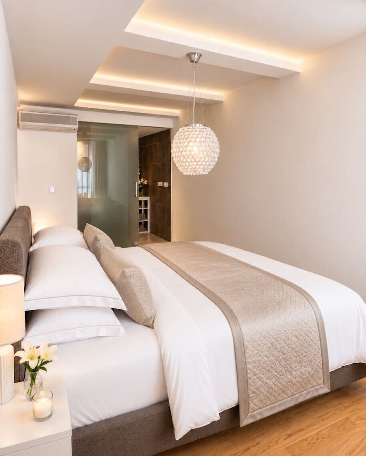A modern bedroom with a large bed, bedside tables, a hanging light fixture, and a view into another room.