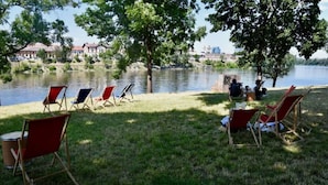 A lakeside area with lawn chairs and a picnic table, a small building, and a view of a cityscape across the water. - Riverside Urban Oasis in Prague’s Coolest District (Prague 8)
