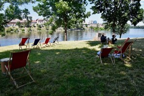 Ein Seeufer mit Liegestühlen und einem Picknicktisch, ein kleines Gebäude und Blick auf eine Stadt am anderen Ufer.