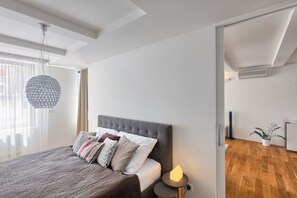 Ein modernes Schlafzimmer mit einem großen Bett, einer Pendelleuchte und Blick in einen angrenzenden Raum.