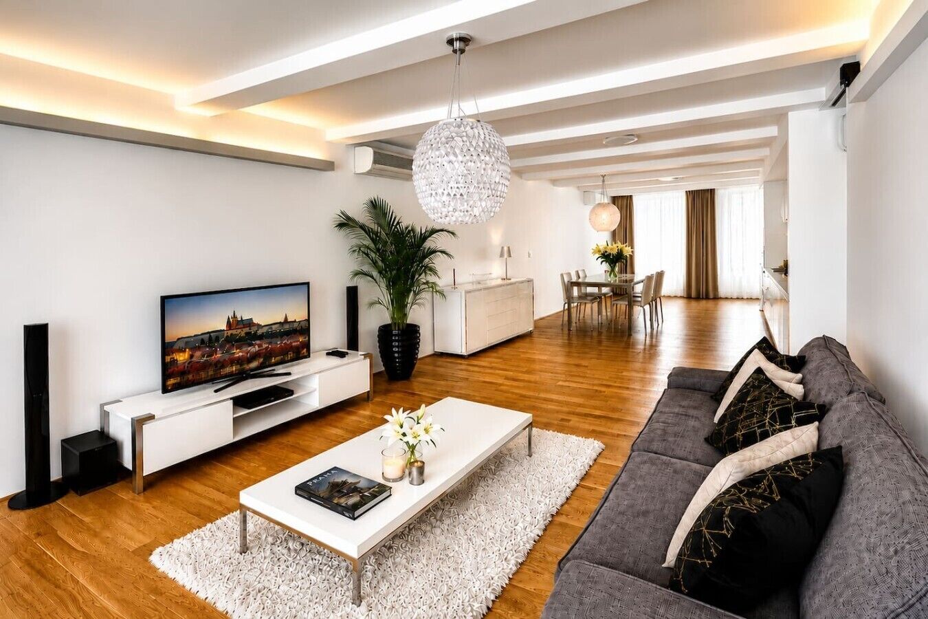 Een moderne woonkamer met een flatscreen televisie, een witte salontafel, een grijze bank en een eethoek op de achtergrond.