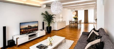 A modern living room with a flat-screen TV, a white coffee table, a grey sofa, and a dining area in the background.