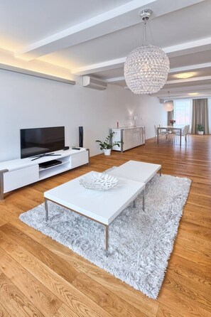 Ein modernes Wohnzimmer mit Flachbildfernseher, weißen Möbeln, einer Kronleuchter und einem Essbereich.
