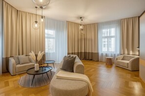 Ein geräumiges Wohnzimmer mit einer Couch, einem Hocker und zwei Beistelltischen, großen Fenstern mit Vorhängen und Holzboden.