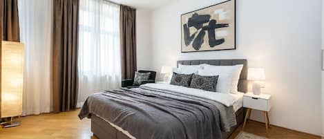 A modern bedroom with a large bed, a bedside table, a lamp, and a window with curtains.