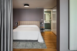 Ein modernes Hotelzimmer mit einem großen Bett, einem Nachttisch, einem kleinen Tisch und einem durchgehenden Türspalt einsehbaren Badezimmer.