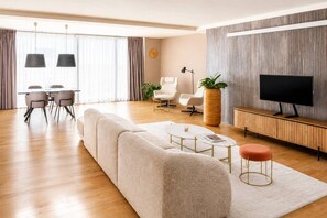 A modern living room with a sectional sofa, a dining table with chairs, a TV, and a large potted plant. - Urban Oasis Karlín – Spacious Retreat by the River (Prague 8)