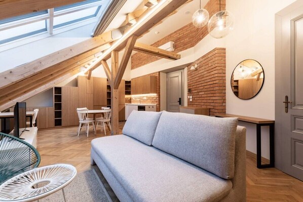 A loft-style room with a high ceiling, exposed wooden beams, a dining area, a sofa, and a kitchen with brick walls. - Elegant Attic Loft in Prime Vinohrady (Prague 2)