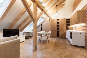 A modern loft with exposed wooden beams, a dining area with white chairs, and a kitchen with wooden cabinetry. - Elegant Attic Loft in Prime Vinohrady (Prague 2)