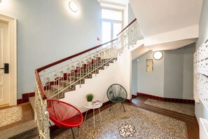 A staircase with ornate metal railings, a red chair, a small table with a plant, and patterned floor tiles. - Cozy Attic Loft in Prime Location (Prague 2)