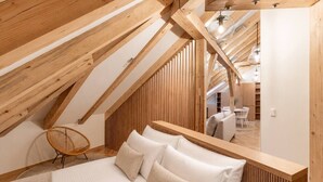 A high-ceilinged attic bedroom with a wooden slat wall, a bed with white linens, a wicker chair, and a view into the dining area. - Cozy Attic Loft in Prime Location (Prague 2)