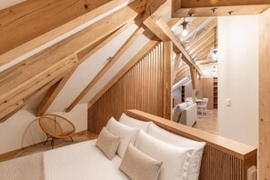 A high-ceilinged attic bedroom with a wooden slat wall, a bed with white linens, a wicker chair, and a view into the dining area. - Cozy Attic Loft in Prime Location (Prague 2)