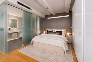Ein modernes Schlafzimmer mit einem großen Bett, Nachttischen mit Lampen, einer Glastrennwand und einem Badezimmer mit Waschtisch und Spiegel.