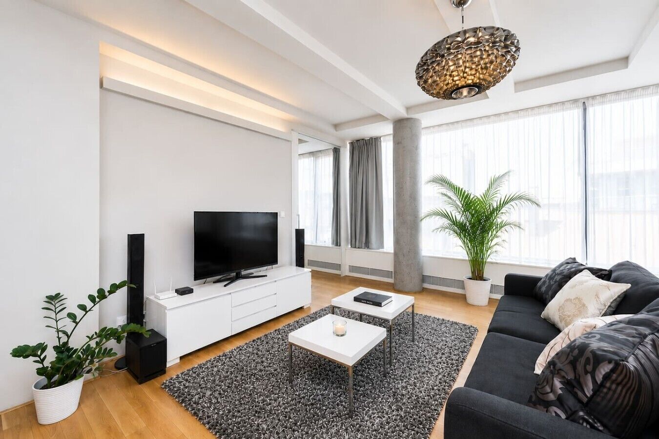 A modern living room with a flat-screen TV, a white entertainment unit, a black sofa, a coffee table, and a large window with curtains.
