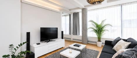 A modern living room with a flat-screen TV, a white entertainment unit, a black sofa, a coffee table, and a large window with curtains.