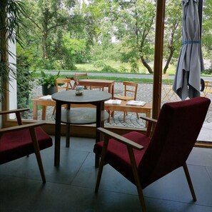 Ein Sitzbereich im Innenbereich mit Blick auf einen Garten. Es gibt Holztische und Stühle, einen kleinen runden Tisch mit Kissen und einen Blumentopf.