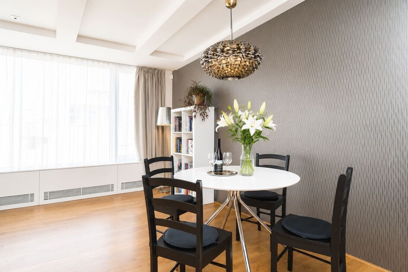 A dining area with a round table, four chairs, a vase of flowers, and a bookshelf.