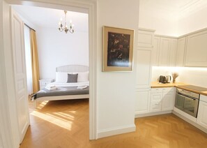 Ein Schlafzimmer mit Bett, Nachttisch und einer Ablage. Es gibt eine Küche mit Schränken, einer Arbeitsplatte und einem Backofen. Ein gerahmtes Bild hängt an der Wand.