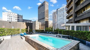 Una piscina en la azotea con terraza de madera, rodeada de edificios de la ciudad.