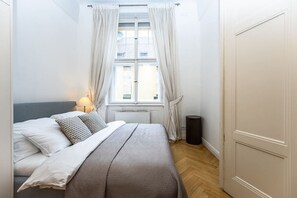 Ein Schlafzimmer mit Bett, Fenster mit Vorhängen, Heizkörper und Mülleimer.