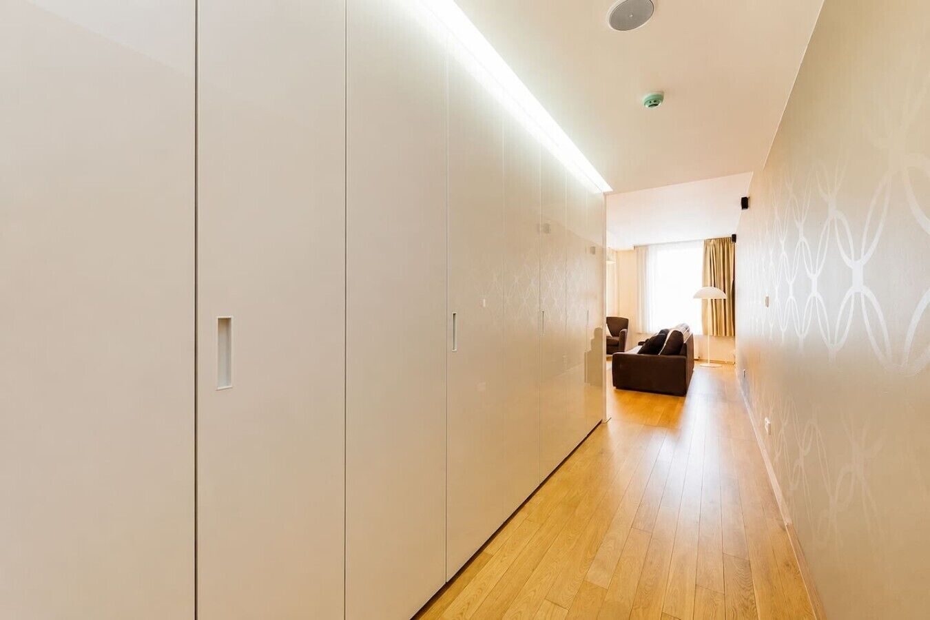 A corridor with a wooden floor leading to a seating area with a sofa and a lamp.