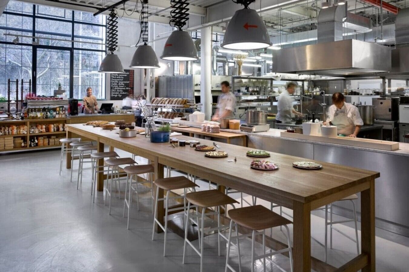 A modern kitchen with a long wooden table, bar stools, hanging pendant lights, and stainless steel appliances.