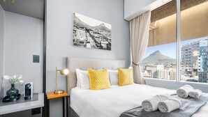 A hotel room with a bed, a desk, a lamp, a vase with flowers, and a cityscape view. - One Thibault 1609 (Cape Town)