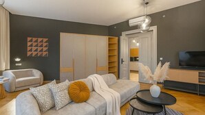 A modern living room with a grey sofa, a coffee table, and a TV. There is a wardrobe and a kitchen visible in the background.