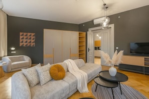 A modern living room with a grey sofa, a coffee table, and a TV. There is a wardrobe and a kitchen visible in the background. - Chic Two-Bedroom Residence in Vinohrady (Praha 2)