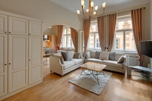 Ein geräumiges Wohnzimmer mit einer Sofagarnitur, einem Couchtisch und einem Fernseher. Im Hintergrund ist eine Küche zu sehen, und das Zimmer verfügt über große Fenster mit Vorhängen.