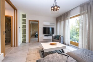Ein modernes Wohnzimmer mit Flachbildfernseher, einer Couch, einem Couchtisch und einem großen Fenster mit Vorhängen.