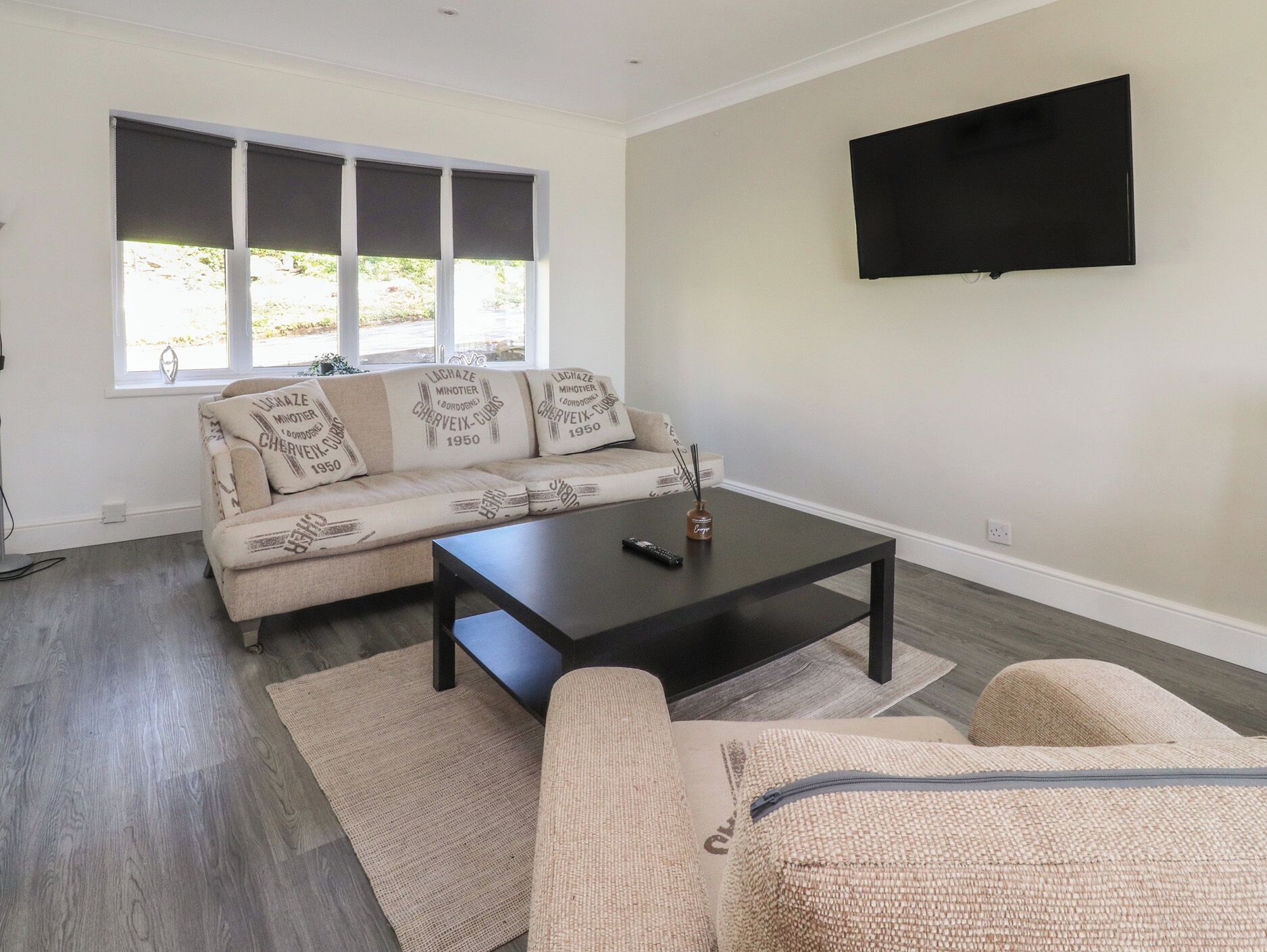 A living room with a sofa, a coffee table, a TV mounted on the wall, and a window with blinds.