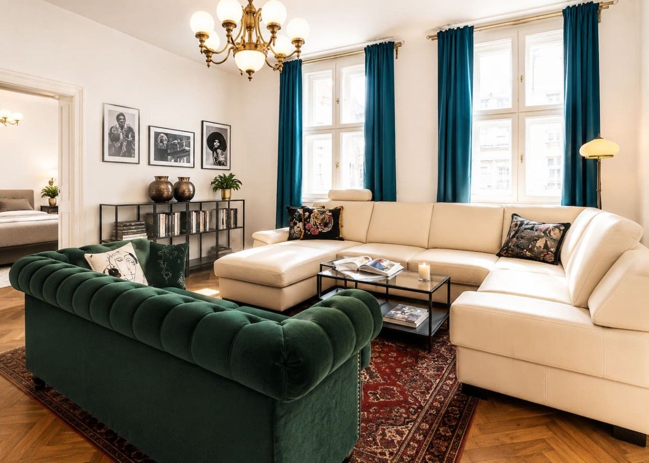 Ein Wohnzimmer mit einem grünen Sofa, einem weißen Sofa und einem Couchtisch. Vorhanden sind Vorhänge, eine Kristallleuchter und gerahmte Bilder an der Wand.