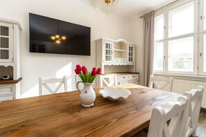 Ein Essbereich mit einem Holztisch, weißen Stühlen, einer weißen Vase mit roten Blumen, einer weißen Schüssel und einem weißen Schrank mit einem an der Wand montierten Fernseher.