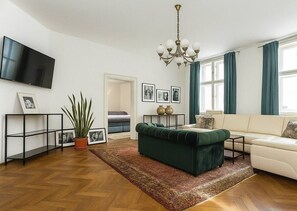 Ein Wohnzimmer mit Flachbildfernseher, einem grünen Sofa, einem weißen Sofa, einer Kristallleuchter und einem großen Fenster mit Vorhängen.
