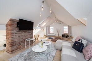 A loft-style living room with a brick fireplace, a flat-screen TV mounted on the wall, a white round coffee table, a beige sofa, and a large window providing natural light. - Old Town 2-BR Loft with Panoramic Prague Castle Views (Praha 1)