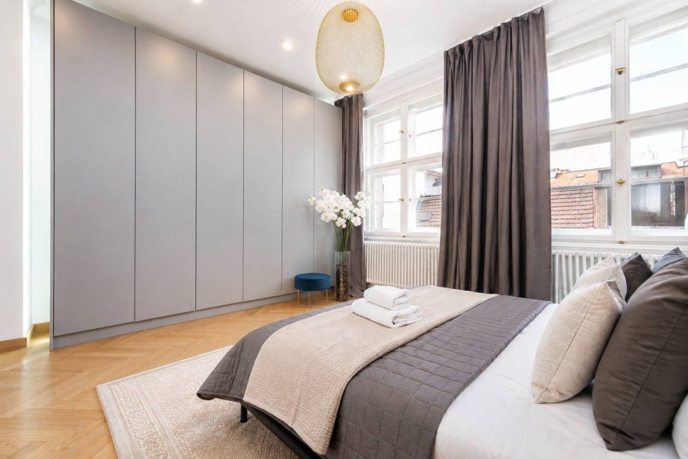 A modern bedroom with a large bed, a bedside table, a vase with flowers, and a view of the city through large windows.