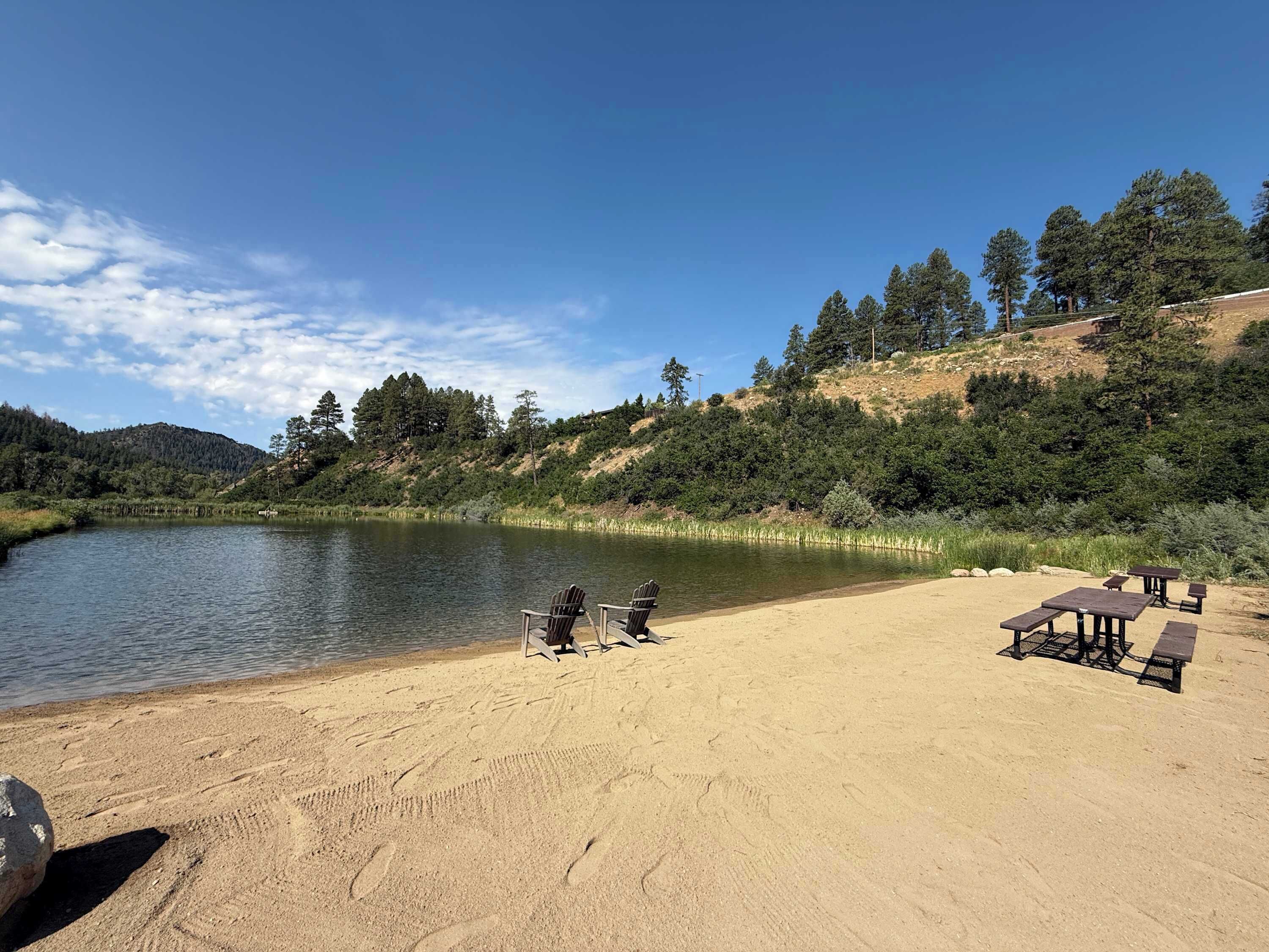 A lakeside beach with picnic tables and chairs, surrounded by forested hills.
