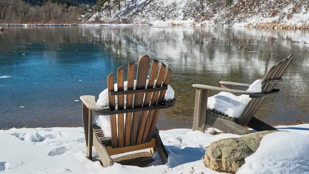 Uma cadeira Adirondack de madeira à beira de um lago congelado, com chão coberto de neve e uma montanha ao fundo.