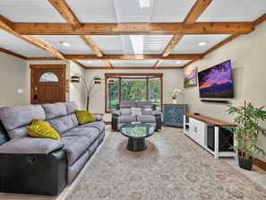Ein Wohnzimmer mit grauem Sofa, Hocker in Schwarz, einem Glastisch, einer blauen Kommode und einer Topfpflanze.