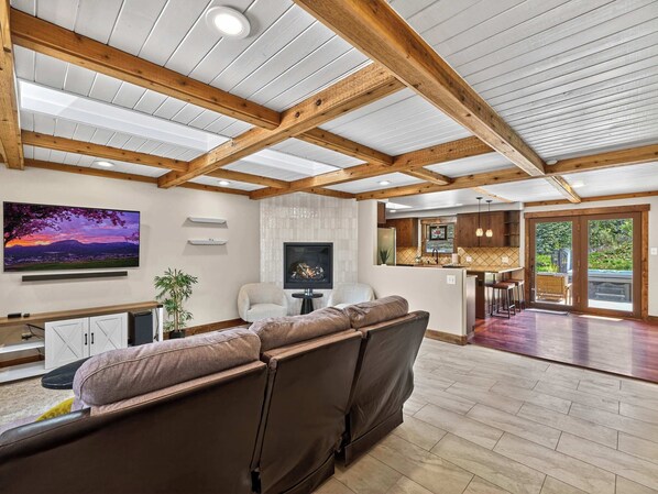 A spacious living area with a large sofa, a flat-screen TV, a fireplace, and a kitchen in the background. - Eagle Ridge River Retreat - Hot Tub, Sauna, Yard! (Durango)