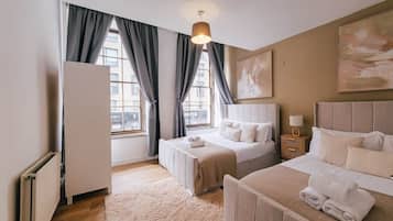 A hotel room with two beds, a radiator, a window with curtains, a painting on the wall, and a bedside table with a lamp.