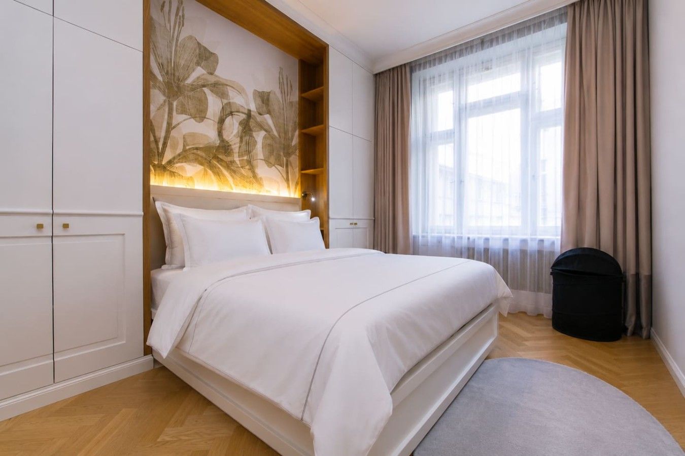 Een hotelkamer met een groot bed, een hoofdbord met een bloemenmotief, een raam met gordijnen en een prullenbak.