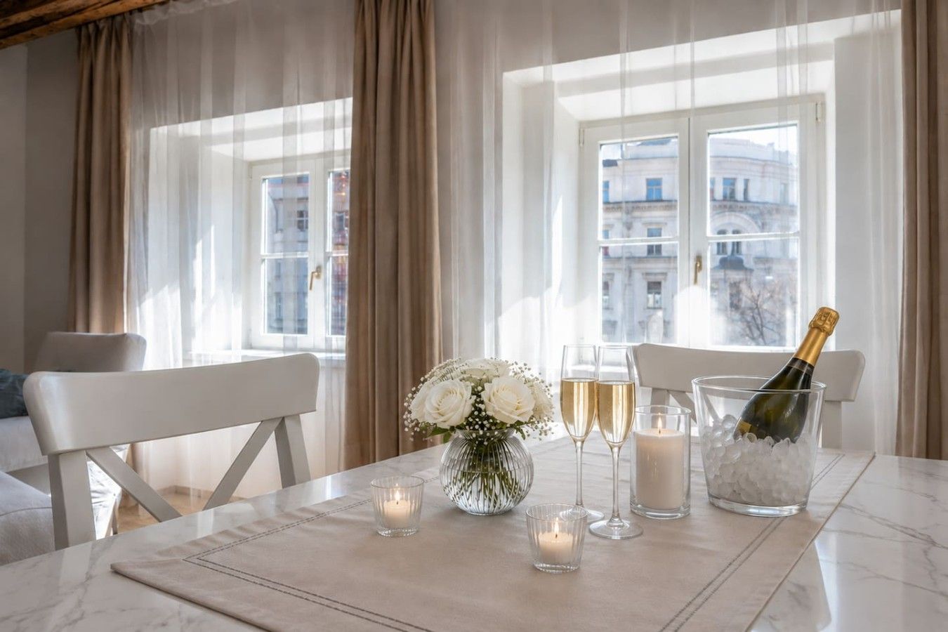 Een eettafel met een vaas witte bloemen, twee champagneglazen, een kaars en een champagnefles in een glazen schaal.